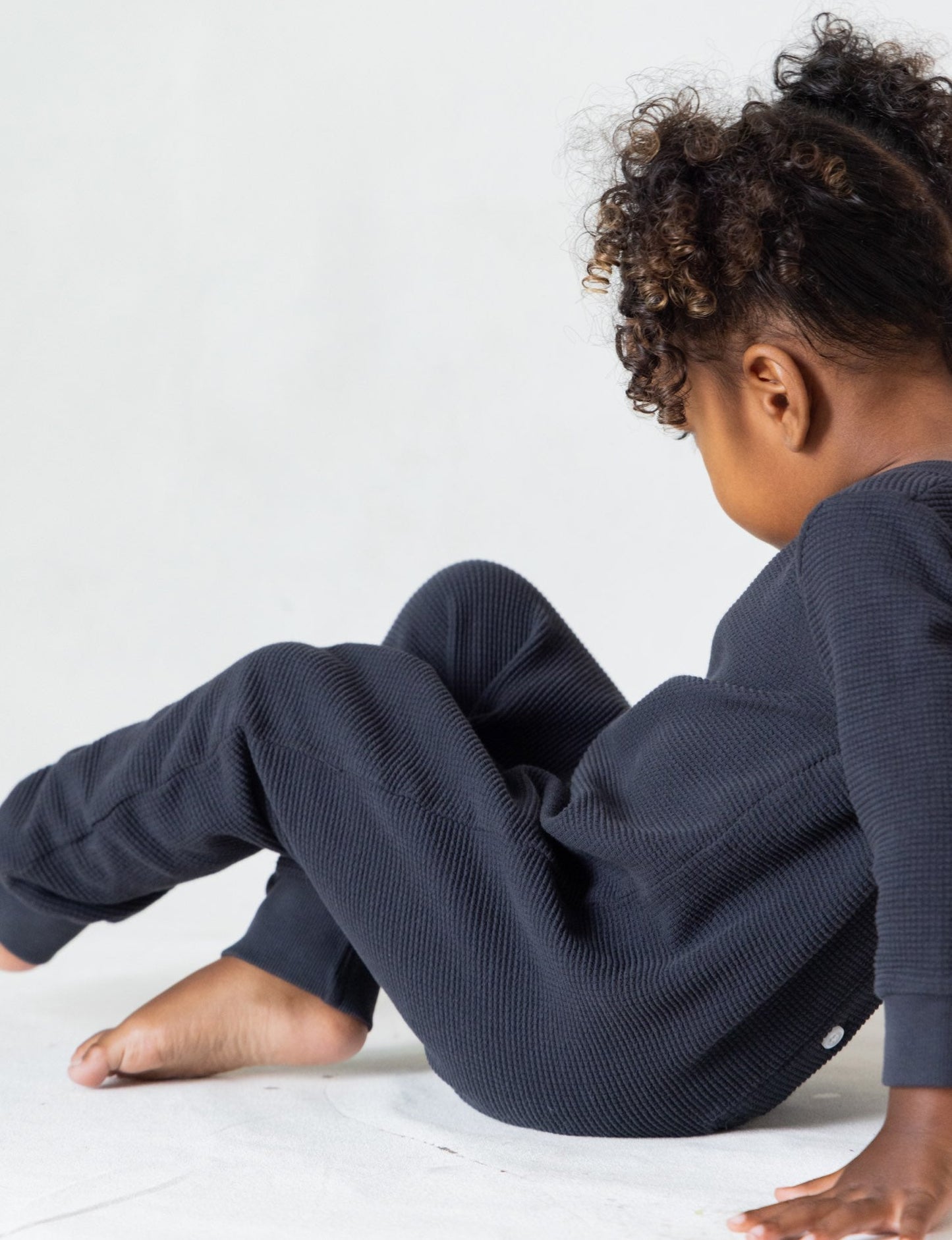 A young child with curly hair sits on the floor, facing away from the camera. They are dressed in The Thermal Onesie by all the babies, a dark-colored thermal outfit, and have bare feet. The background is plain and light-colored.