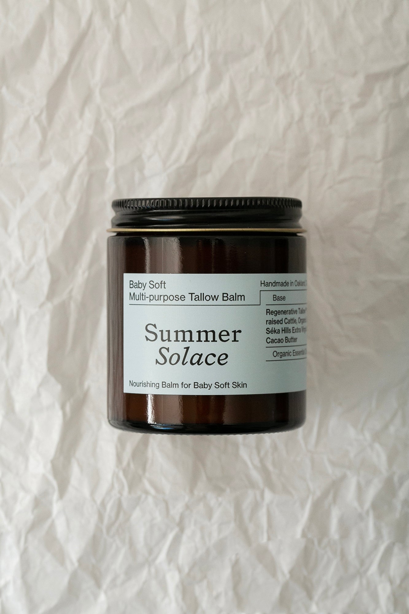 A brown glass jar with a black lid, labeled "Summer Solace Tallow," contains "Baby Soft Family Balm - Regenerative Tallow™ + Pastured Leaf Lard." This nourishing moisturizer is perfect for baby soft and sensitive skin. The label highlights that it is handmade in Oakland and rich in organic fats and essential oils.