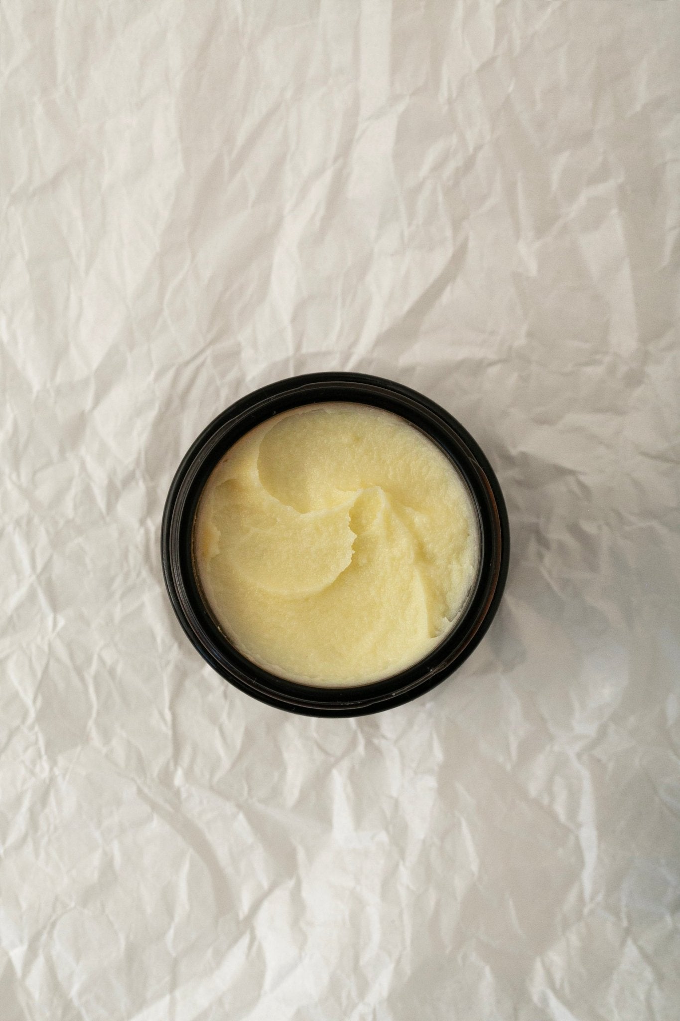 A small black jar labeled "Baby Soft Family Balm - Regenerative Tallow™ + Pastured Leaf Lard" by Summer Solace Tallow rests on a crumpled white paper background. The creamy, off-white baby moisturizer inside appears smooth and whipped, making it ideal for sensitive skin.