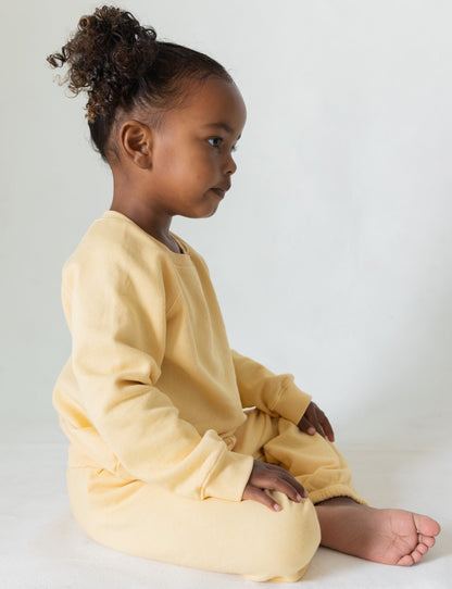 A young child with curly hair sits cross-legged on the floor, wearing The Lil' Classic Sweatpant from all the babies, crafted from organic French terry cloth. The matching light yellow sweatshirt and vintage-inspired sweatpants enhance their serene expression against the plain, light-colored background that focuses attention on their side profile.