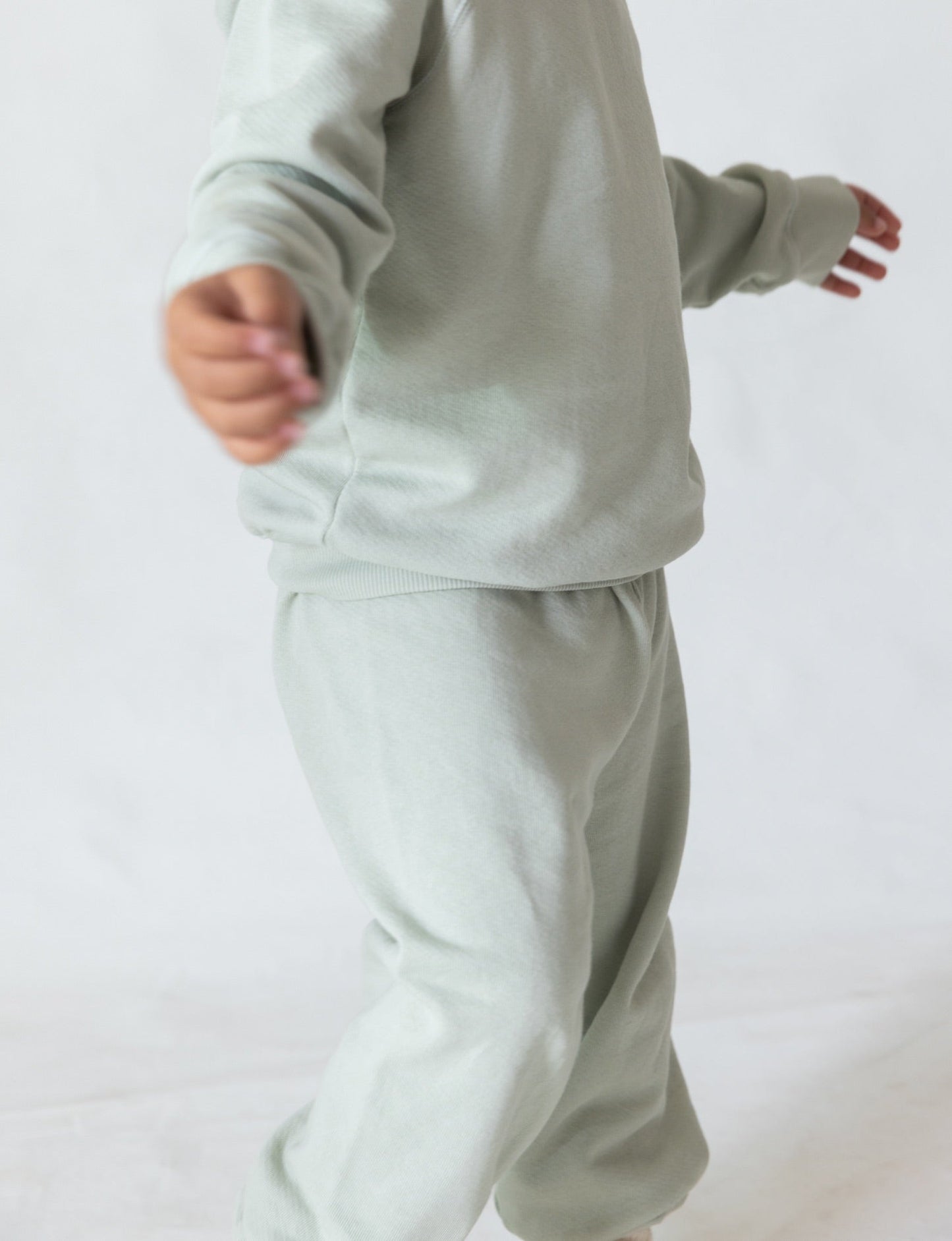 A person dressed in The Lil' Classic Crewneck by all the babies and matching sweatpants made of organic French terry cloth extends their arms outward and slightly away from the camera. The background is plain and white, creating a minimalist aesthetic.