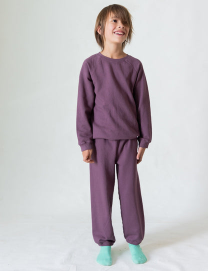 A young child stands against a plain background, smiling and wearing matching purple long-sleeved sweatshirt and The Lil' Classic Sweatpant from all the babies, crafted from organic French terry cloth. The child also has on light blue socks. The overall look is relaxed, cozy, and comfortable.