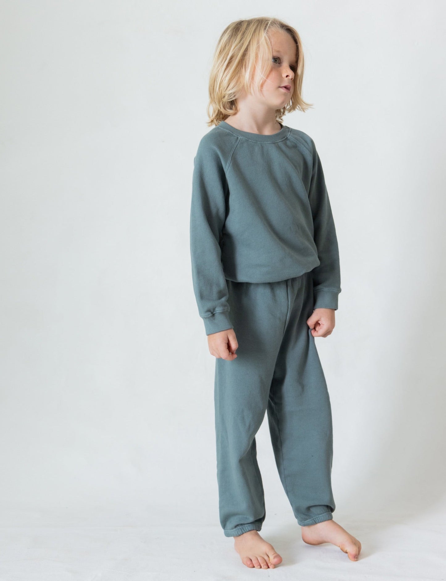 A young child with shoulder-length blonde hair stands barefoot against a plain background, dressed in The Lil' Classic Sweatpant by all the babies, made from cozy organic French terry cloth. The light blue sweatshirt and vintage-inspired sweatpants set complement the child's relaxed posture as they gaze off to the side.