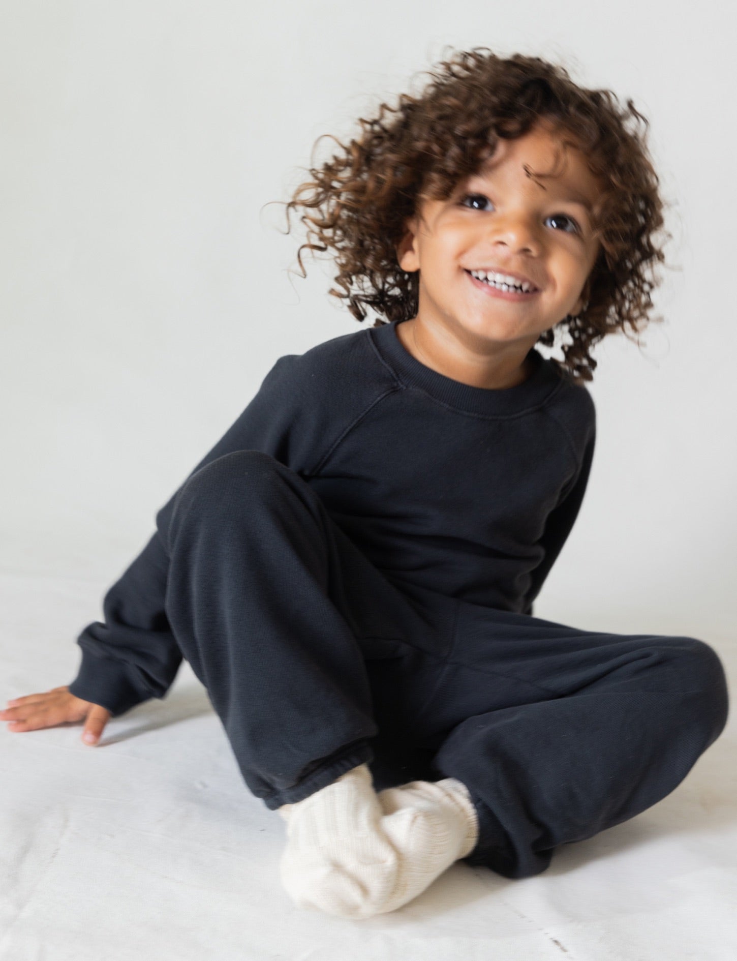 A young child with curly hair smiles while sitting on the floor, dressed in a cozy, dark long-sleeved outfit called The Lil' Classic Sweatpant by all the babies and white socks made of organic French terry cloth. The background is plain and light-colored.