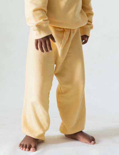 A young child stands barefoot on a white surface, wearing an oversized pale yellow sweat suit made from organic French terry cloth. The outfit includes a sweatshirt and The Lil' Classic Sweatpant by all the babies, slightly too big for the child, creating a cozy and relaxed appearance. The child's hands and feet are visible.