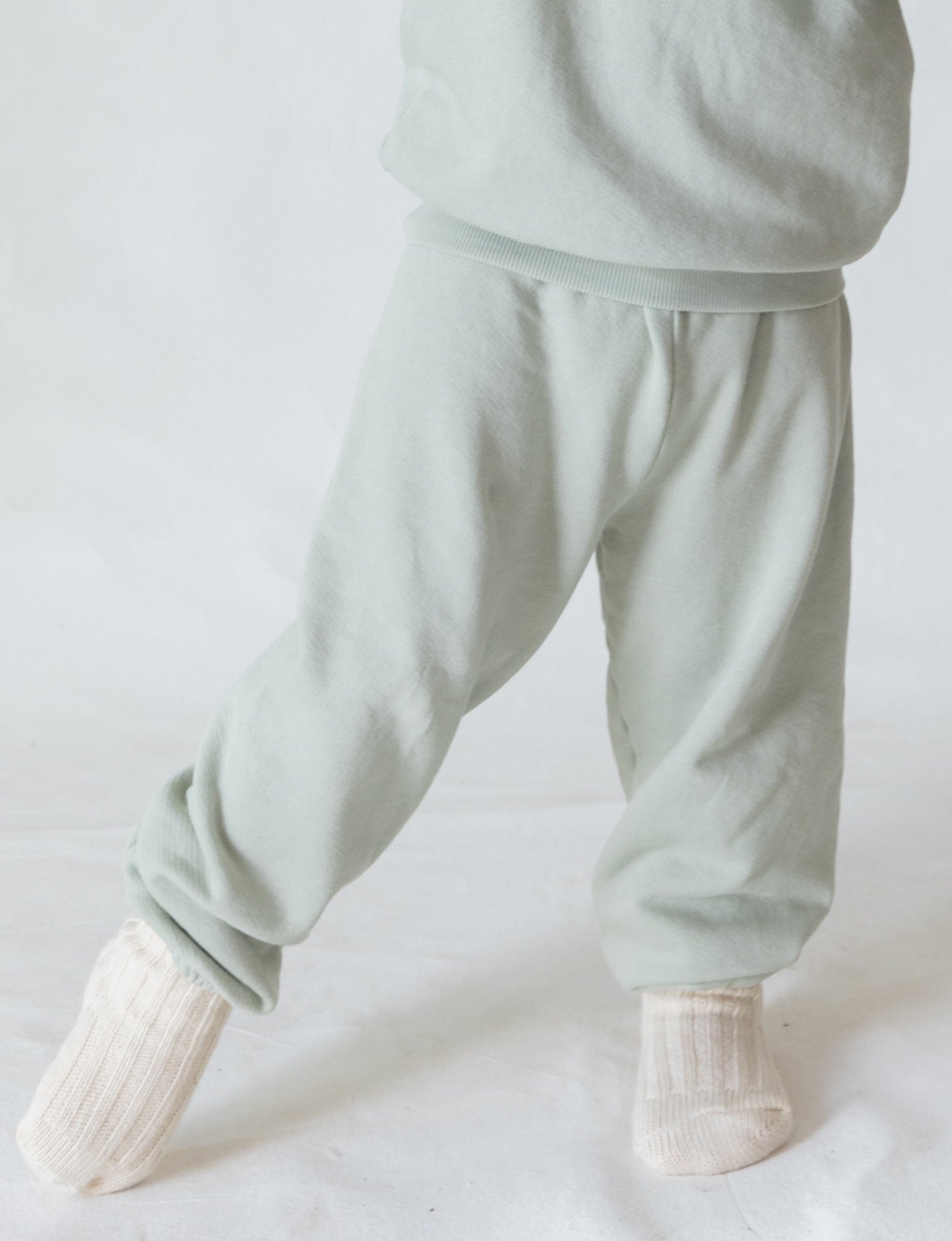 A child wearing The Lil' Classic Sweatpant in light green by all the babies stands with their legs slightly apart, facing forward. The child's feet are clad in soft, white, knitted socks, and their upper body is not visible in the image. The background is plain white.