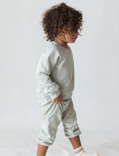 A young child with curly brown hair is walking to the right. They are dressed in a cozy outfit that includes The Lil' Classic Sweatpant by all the babies, paired with a matching light green long-sleeve top. Both pieces are crafted from organic French terry cloth. The child also wears white socks, and the background is plain and light-colored.