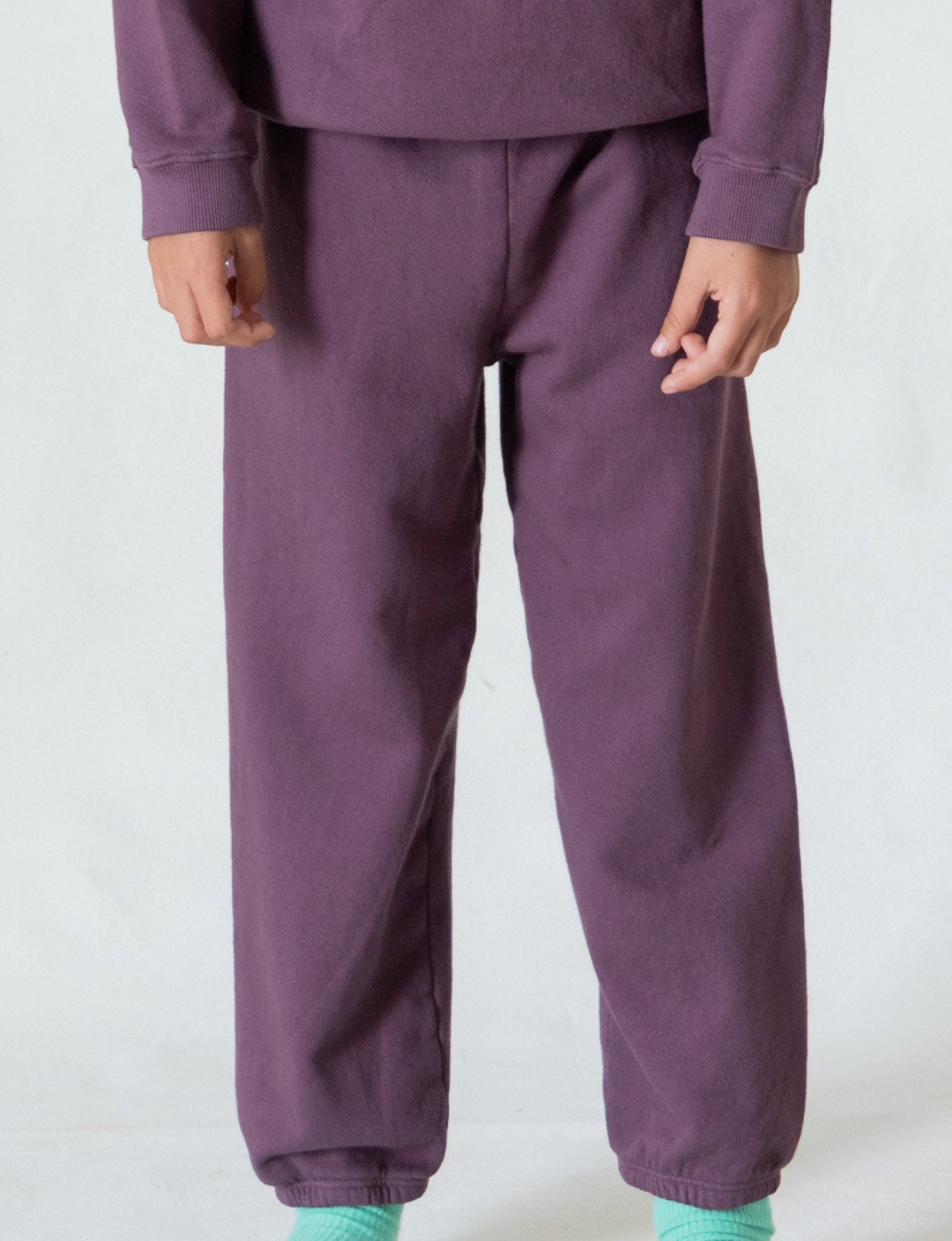 A person wearing *The Lil' Classic Sweatpant* by all the babies in a vintage-inspired dark purple hue, paired with a matching sweatshirt, stands against a plain light background. Only the lower half of their body is visible. They are also sporting mint green socks and no shoes, creating an effortlessly cozy outfit.