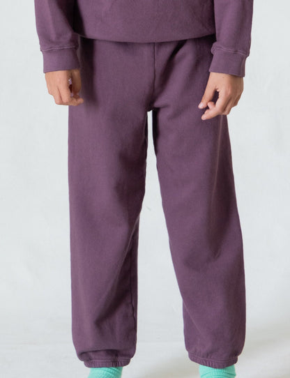 A person wearing *The Lil' Classic Sweatpant* by all the babies in a vintage-inspired dark purple hue, paired with a matching sweatshirt, stands against a plain light background. Only the lower half of their body is visible. They are also sporting mint green socks and no shoes, creating an effortlessly cozy outfit.