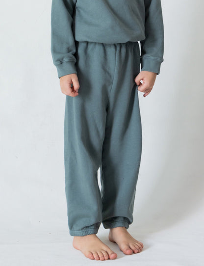 A child stands barefoot against a plain white background, wearing a long-sleeve, teal-colored top and The Lil' Classic Sweatpant by all the babies. The cozy outfit features organic French terry cloth, with the child's hands resting at their sides, exuding casual comfort.