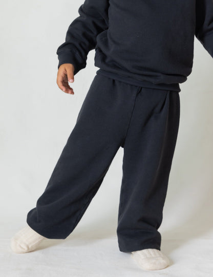 A person wearing a cozy outfit of loose-fitting black clothes, featuring a sweatshirt made from organic French terry cloth and The Lil' Classic Sweatpant by all the babies, stands against a plain white background. Their face is not visible, and they are wearing white socks.