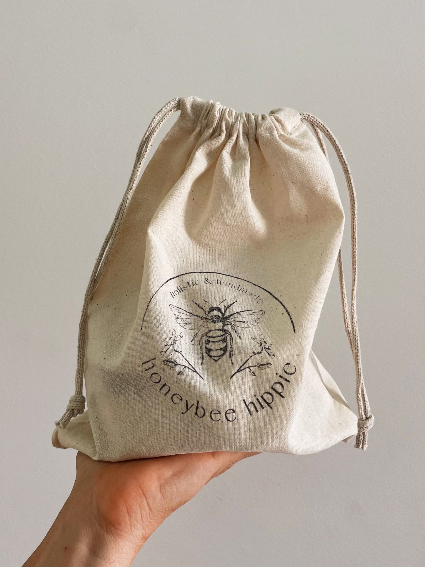 A hand holds a natural fabric drawstring pouch that says "Honeybee Hippie" with a bee illustration, part of the "the honey bundle", and exudes a subtle honey glow against the plain background.