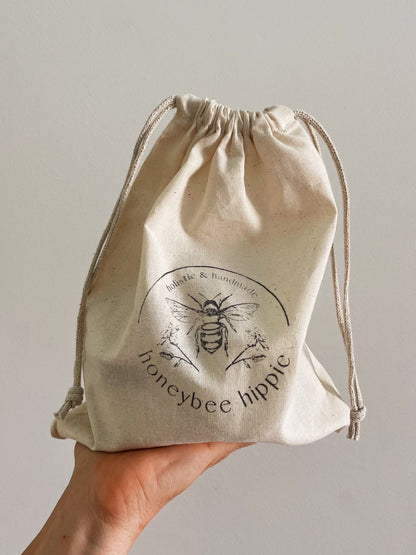 A hand holds a natural fabric drawstring pouch that says "Honeybee Hippie" with a bee illustration, part of the "the honey bundle", and exudes a subtle honey glow against the plain background.