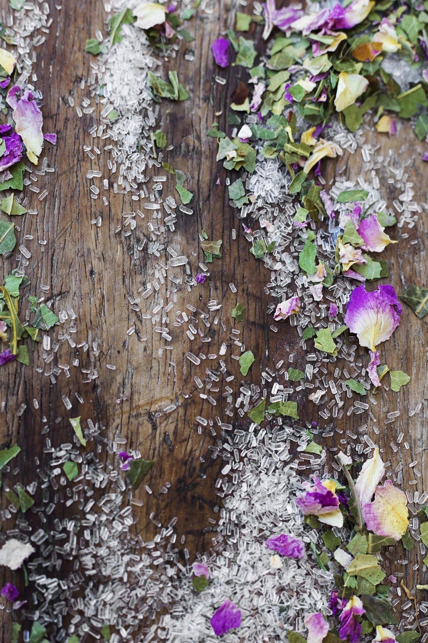 Close-up of a wooden surface scattered with grains of rice, Surya Calming Bath Soak's purple and white flower petals, green leaves, and sprinkles of natural salts. The arrangement appears random, creating a textured and colorful display.