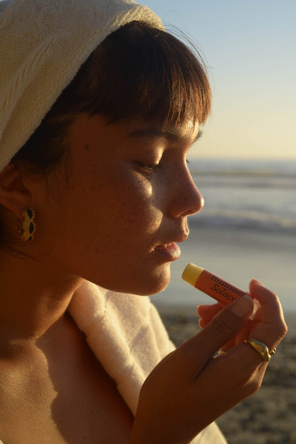 A person with a towel wrapped around their head holds a tube of Summer Solace Tallow's Cardamom & Blood Orange Lip Balm- Regenerative Tallow™ near their lips. They are standing outdoors by the beach at sunset, eyes closed, basking in the warm light. The scent of Italian blood orange mingles with the salty sea air, creating a blissful moment of serenity.