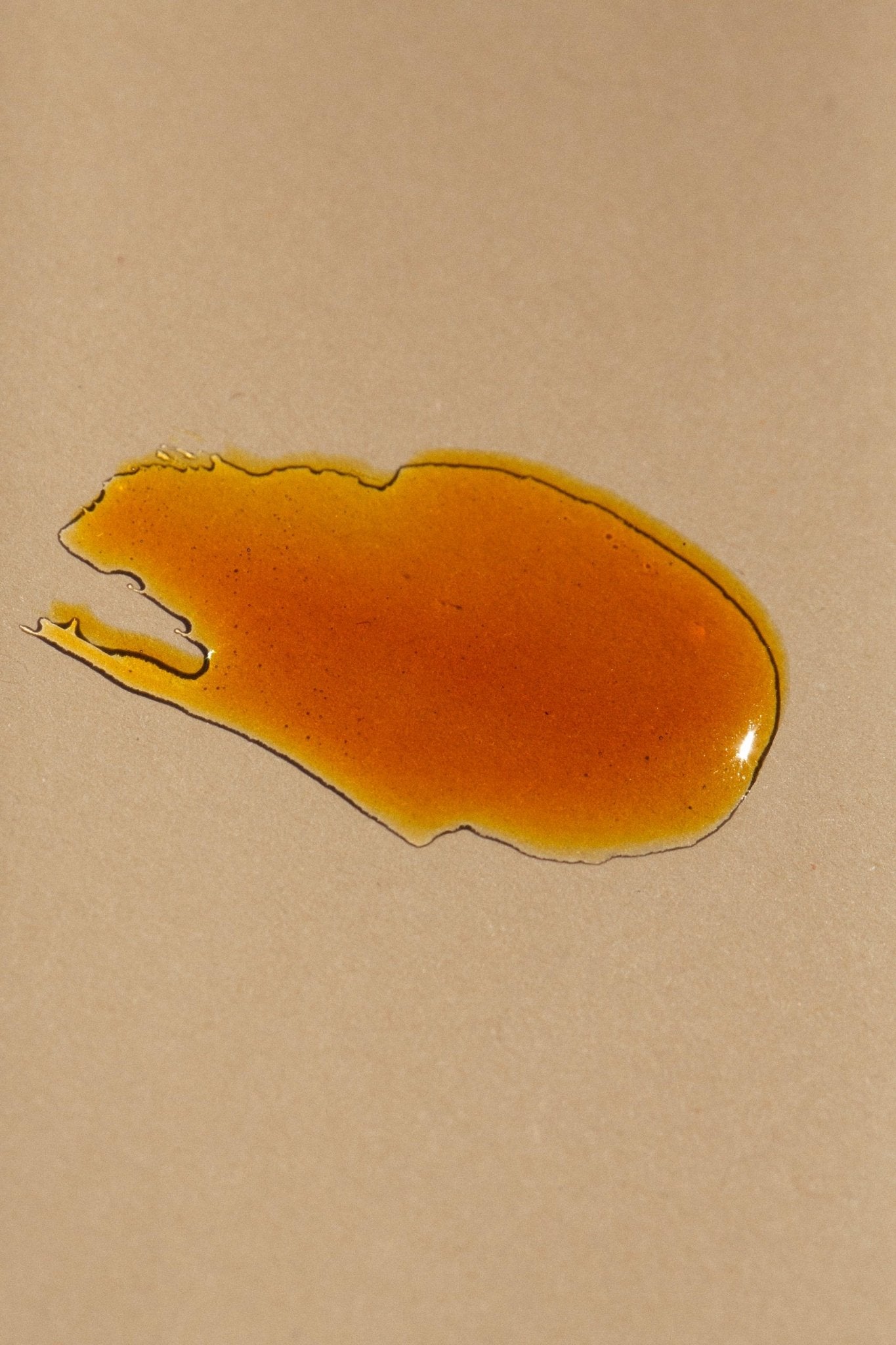 A small, irregularly-shaped puddle of amber-colored liquid, reminiscent of Summer Solace Tallow's Cardamom & Blood Orange Lip Balm- Regenerative Tallow™, is splattered on a smooth, beige surface. The liquid appears glossy and slightly viscous, spreading out unevenly with a few jagged edges.