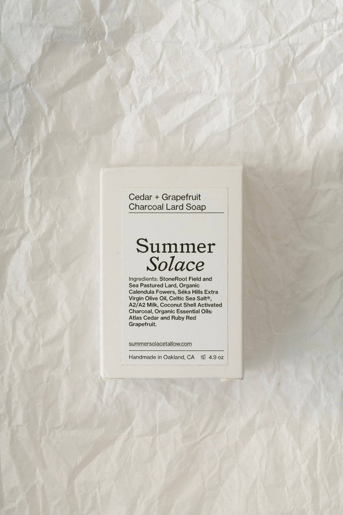 A rectangular bar of Cedar and Grapefruit Charcoal Pastured Leaf-Lard Soap, branded "Summer Solace Tallow," enriched with activated charcoal and essential oils, sits on a crinkled white surface. The label lists ingredients, its handmade origin in Oakland, CA, and its weight of 4.5 oz. The product's website URL is also displayed.