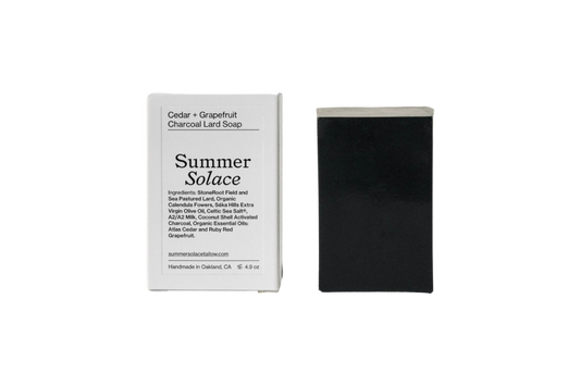 A bar of Summer Solace Tallow Cedar and Grapefruit Charcoal Pastured Leaf-Lard Soap is displayed beside its packaging. The packaging lists the ingredients and essential oils used, including lard, grains, shea, sunflower, castor, avocado, and grapefruit. It mentions that the soap is handmade in Oakland, CA.