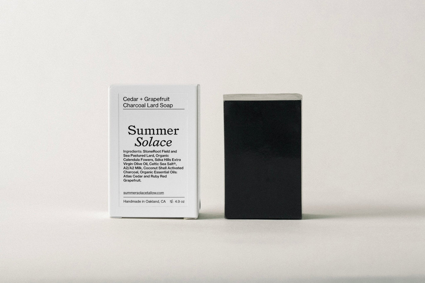 A white box labeled "Summer Solace Tallow" with details about Cedar and Grapefruit Charcoal Pastured Leaf-Lard Soap stands next to a rectangular black soap bar. The box highlights ingredients like activated charcoal, leaf-lard soap, and essential oils; all handmade in Oakland, CA.