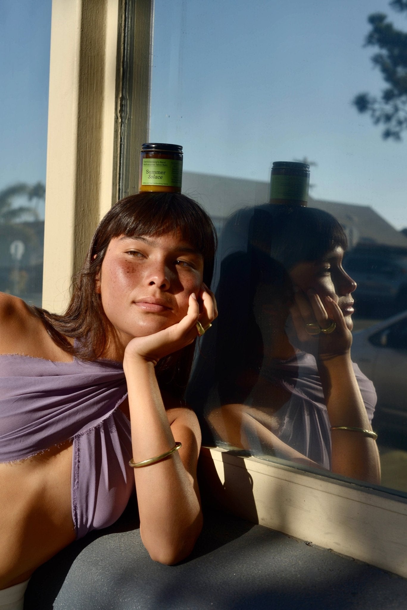 A person with light brown hair and bangs, wearing a lavender top and a gold bracelet, is leaning against a window with a serene expression after using Summer Solace Tallow's Chef & Gardener's Balm - Regenerative Tallow™. There is a jar balanced on their head, and their reflection is visible in the glass. The background reveals an outdoor setting.