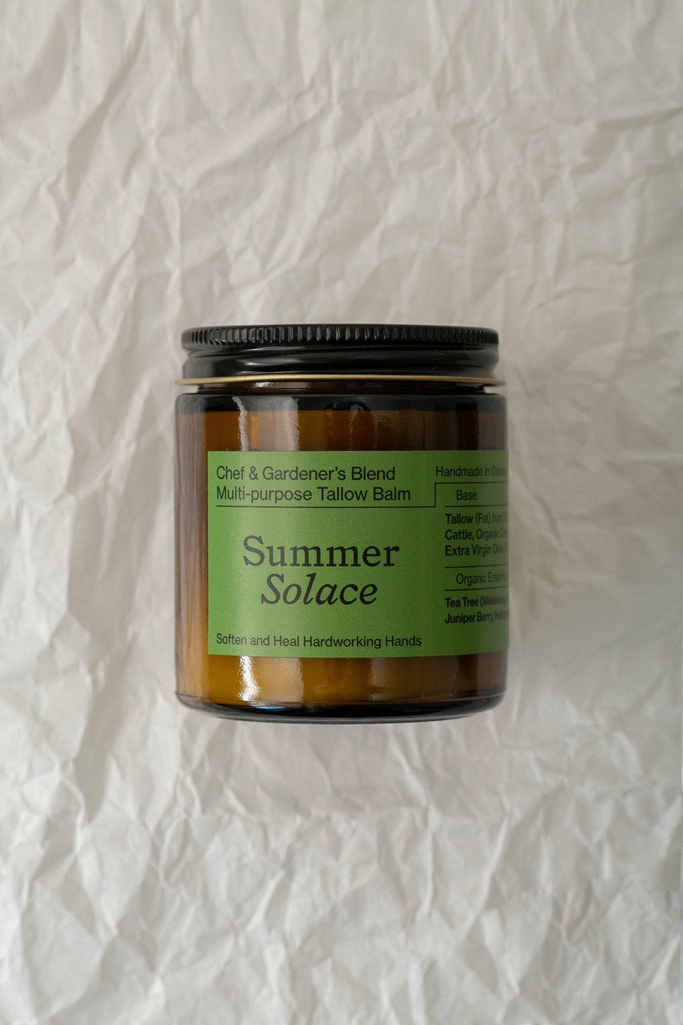 A brown glass jar labeled "Summer Solace Tallow Chef & Gardener's Balm - Regenerative Tallow™" with a black screw lid. The label mentions it's for softening and healing hardworking hands, featuring therapeutic skin treatment ingredients like tallow, olive oil, and healing rosemary and helichrysum. Background is crumpled white paper.