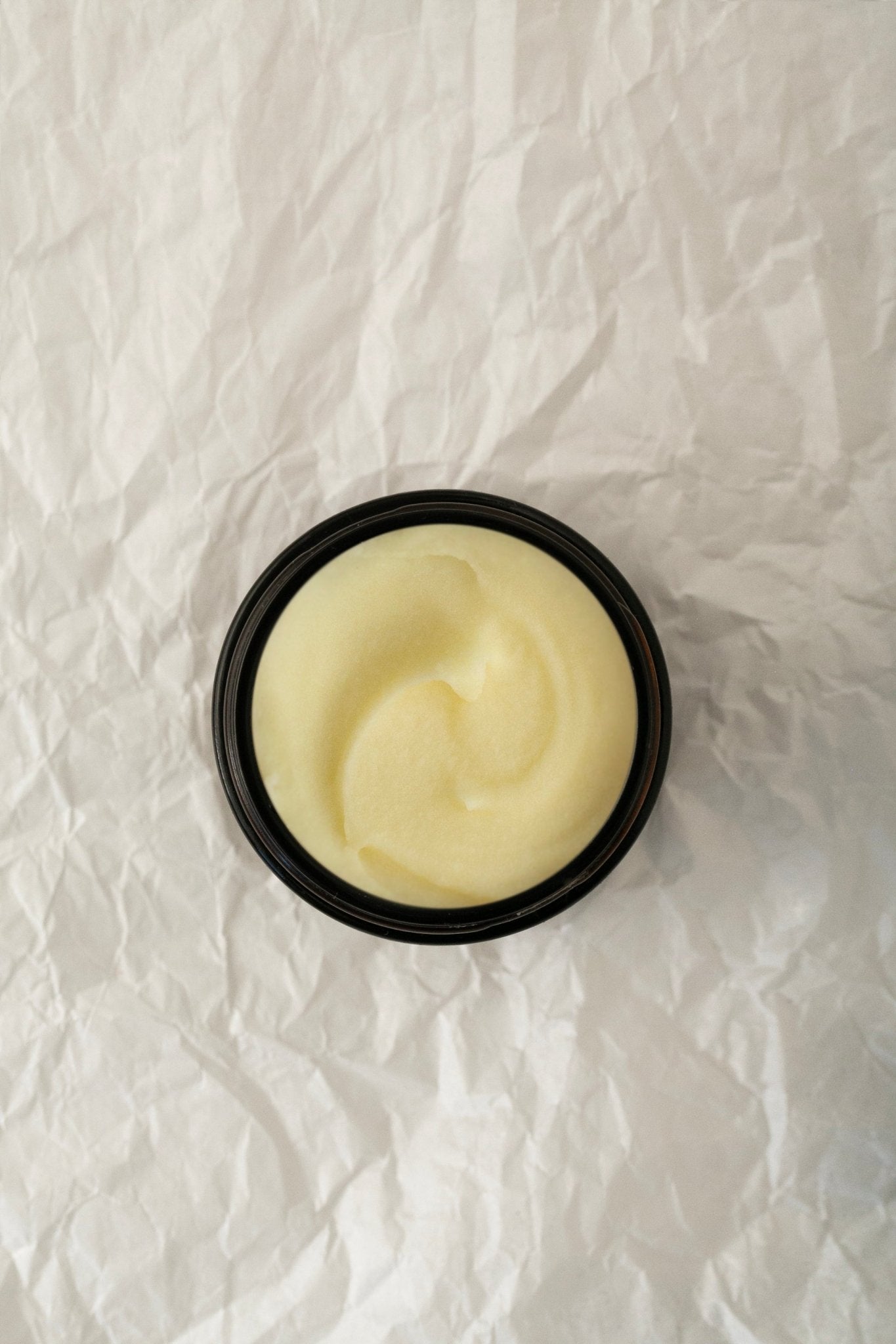 A small, round, black container of Chef & Gardener's Balm - Regenerative Tallow™ by Summer Solace Tallow is placed on a crumpled white paper background. The creamy, light yellow surface of the hydrating tallow balm appears smooth with a subtle swirl pattern in the center.