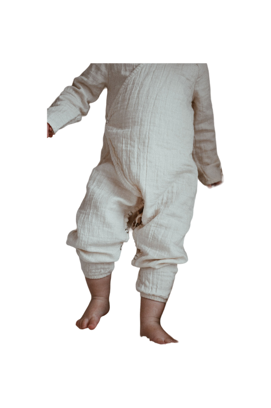 A small child, dressed in the cozy APRÈS WRAP undyed one-piece outfit by cabane childrenswear, stands against a dark background. The organic cotton layette, quilted from neck to ankle, leaves only the child's bare feet and partial upper body visible.