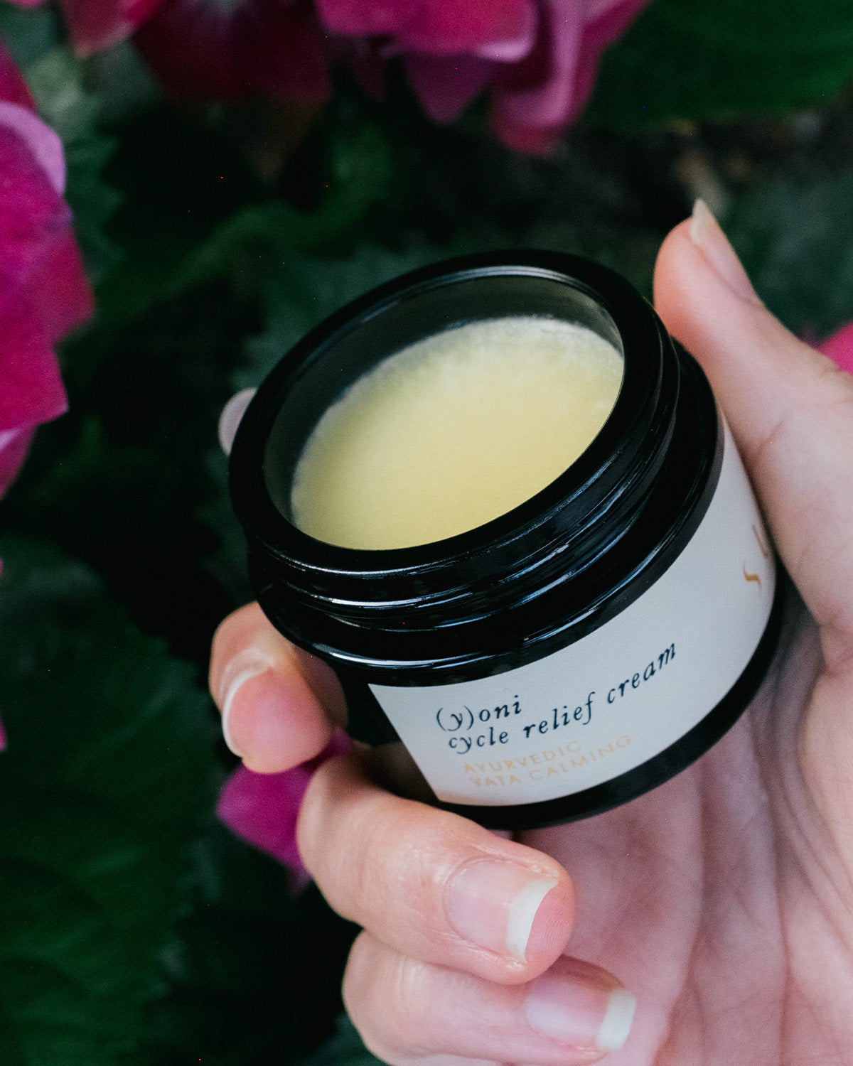 A hand holds an open container of Surya's (y)oni Cycle Relief Cream, infused with Ayurvedic botanicals, against a backdrop of pink flowers and green leaves. The cream inside the container is light yellow, resembling ghee. The label on the jar includes other partially visible text.