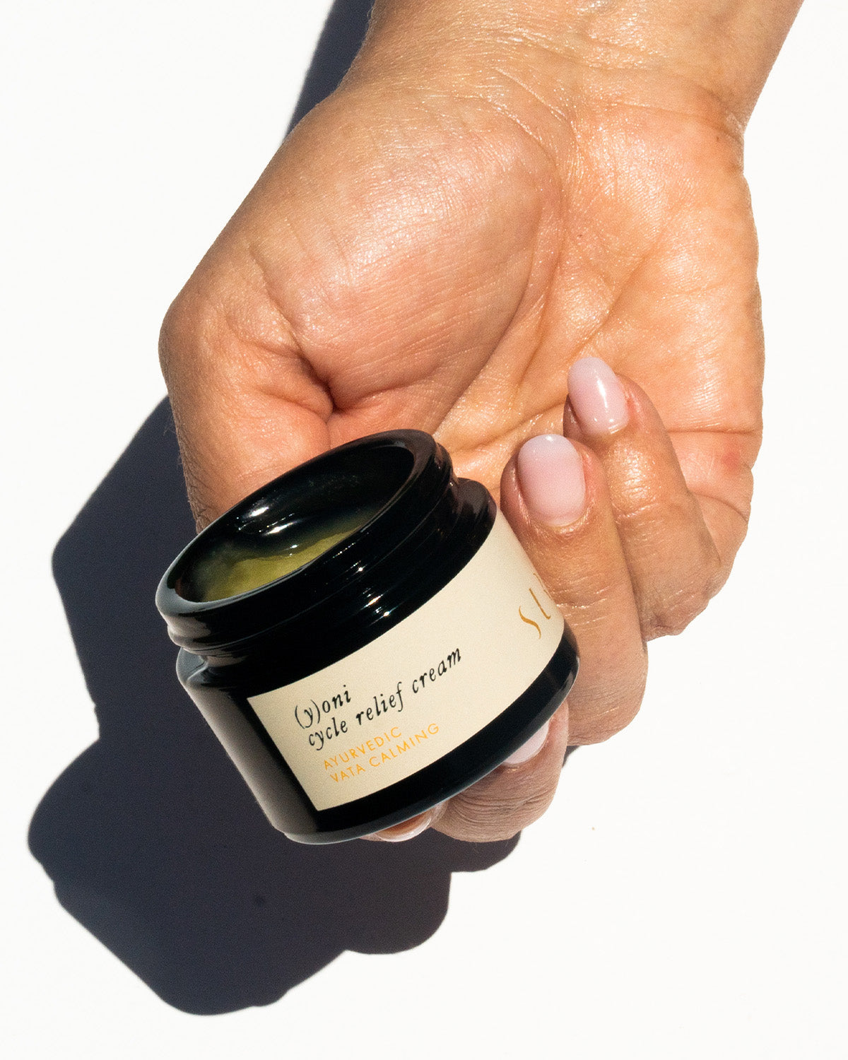 A hand with well-manicured nails holds an open black jar labeled "(y)oni Cycle Relief Cream" from the Surya brand. The person has light skin, and the white background showcases a shadow cast by the hand and jar. The cream, enriched with Ayurvedic botanicals, promises relief for intimate skin during cycles.