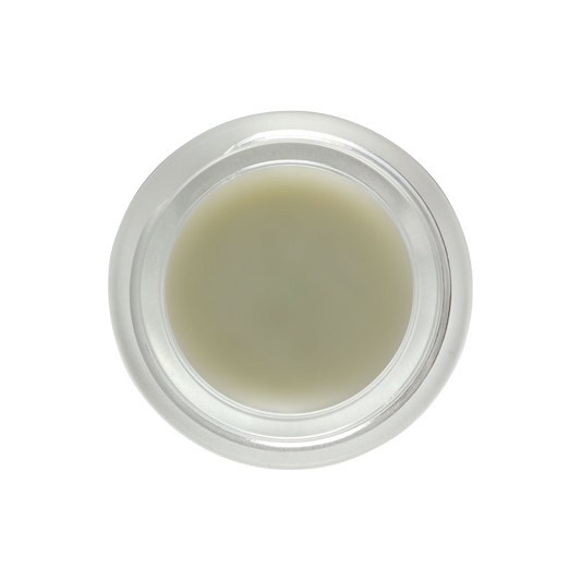 A top-down view of a clear, round container filled with Living Libations' DewDab - Ozonated Beauty Balm in a light beige hue. The container features a smooth, transparent lid. Set against an entirely black background, the container and its pure plant intelligence contents become the focal point of the image.