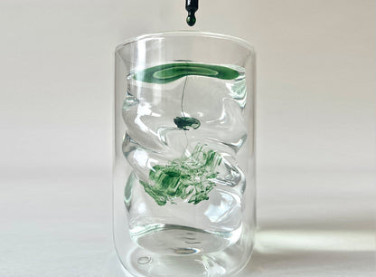 A drop of Juna's Chlorophyll Detox Drops falls into a clear glass of water, symbolizing the benefits of liver detoxification. The drop swirls and disperses, creating abstract, cloud-like patterns in the water. The background is plain and light-colored, drawing attention to the dynamic movement within the glass.