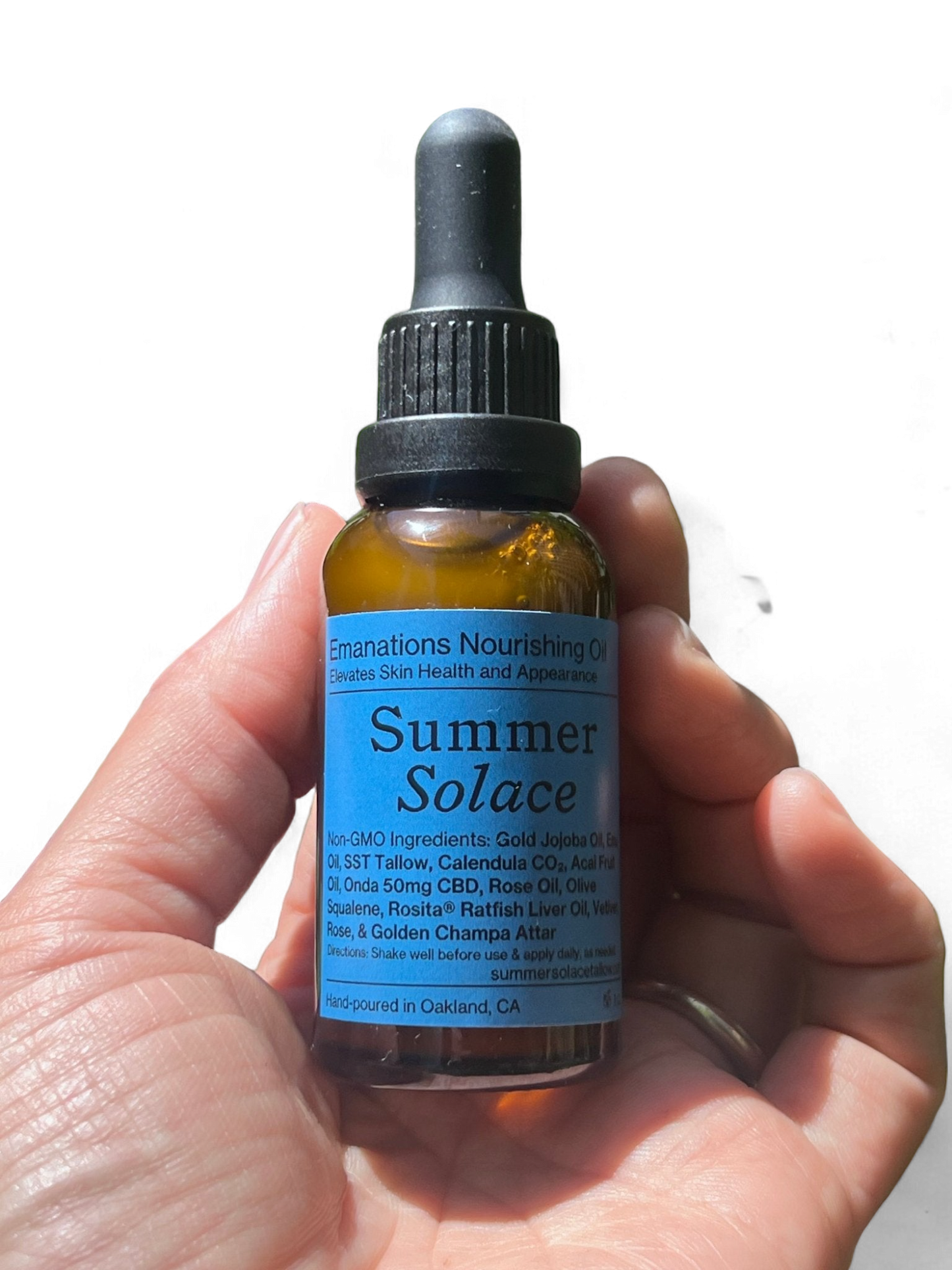 A hand holds a brown dropper bottle labeled "Summer Solace Tallow" with blue text. The label highlights the luxury skincare oil's non-GMO ingredients and benefits for skin health and appearance. Hand-poured in Oakland, CA, this Emanations Nourishing Face and Hair Oil-Animal-based is free from animal-based ingredients.