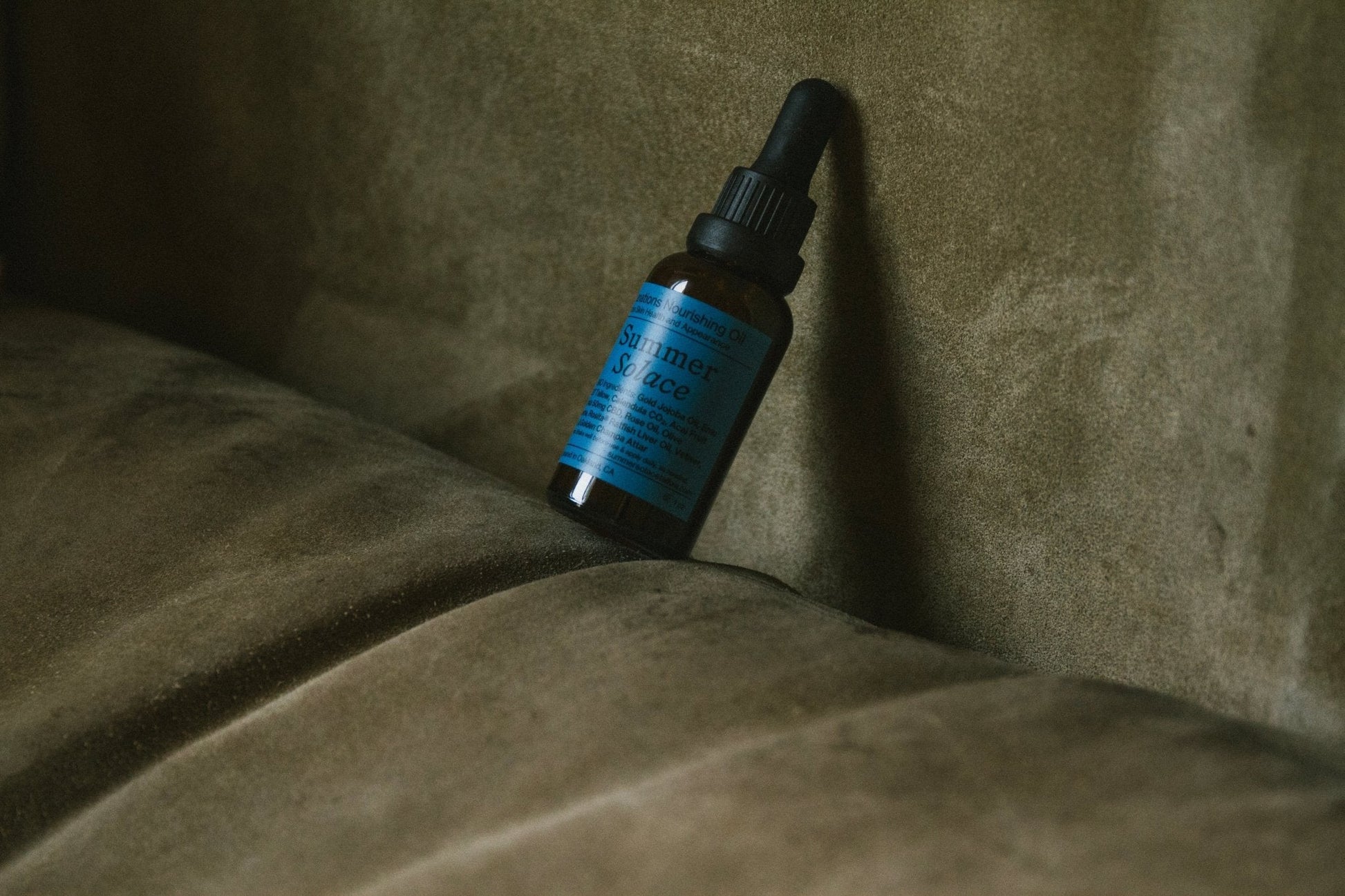 A small brown dropper bottle with a blue label stands upright against the back of a plush, olive green sofa. The text on the label is partially legible and includes "Emanations Nourishing Face and Hair Oil-Animal-based," hinting at luxury skincare from Summer Solace Tallow. The lighting is soft, casting gentle shadows on the sofa's surface.