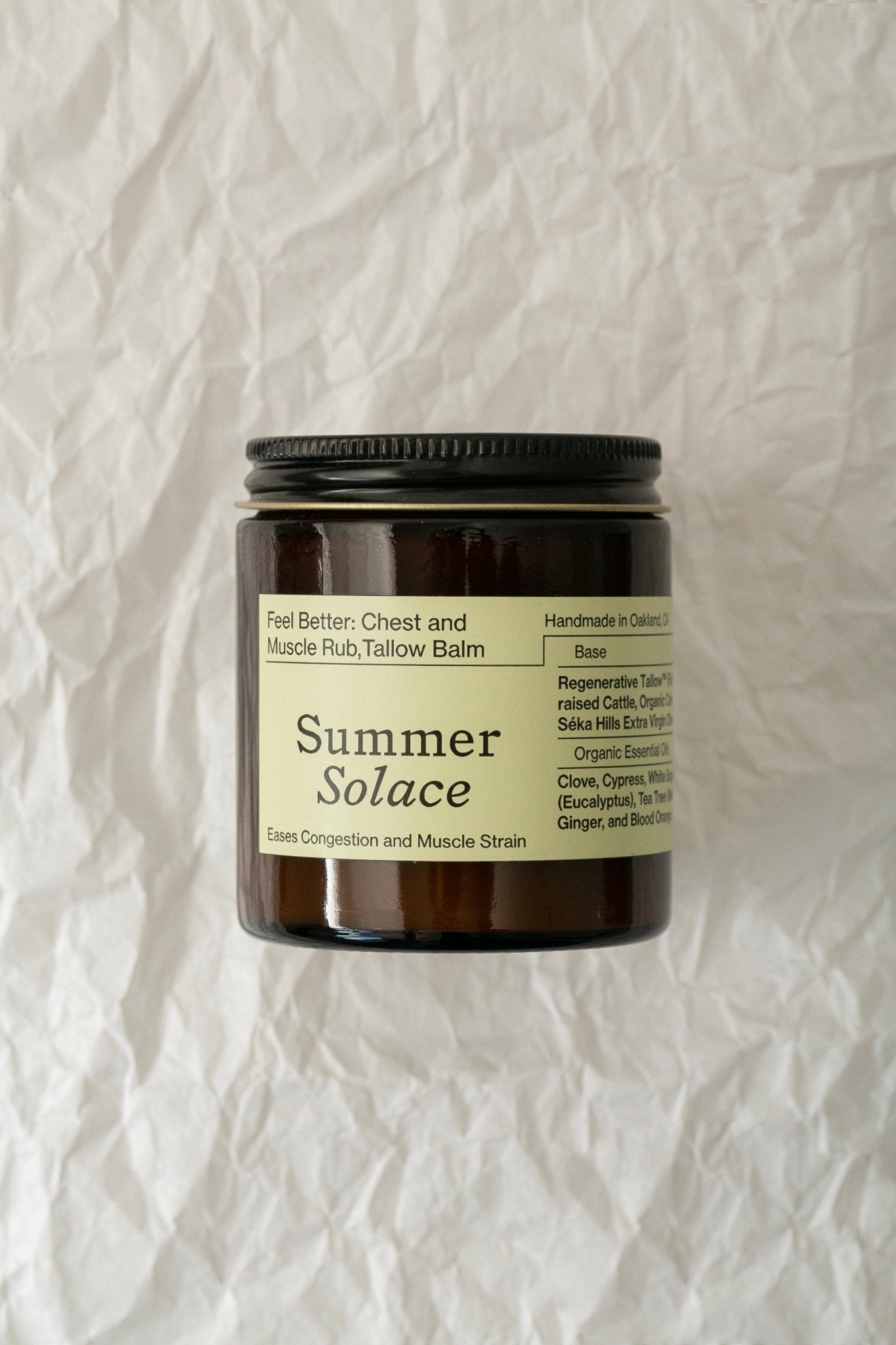 A dark brown jar of Summer Solace Tallow's Feel Better: Chest & Muscle Rub Balm - Regenerative Tallow™ sits on a crinkled white background. The label states it is handmade in Oakland, CA, and lists ingredients including clove, cypress, tallow, eucalyptus, and ginger—perfect for aromatherapy immune soother benefits.