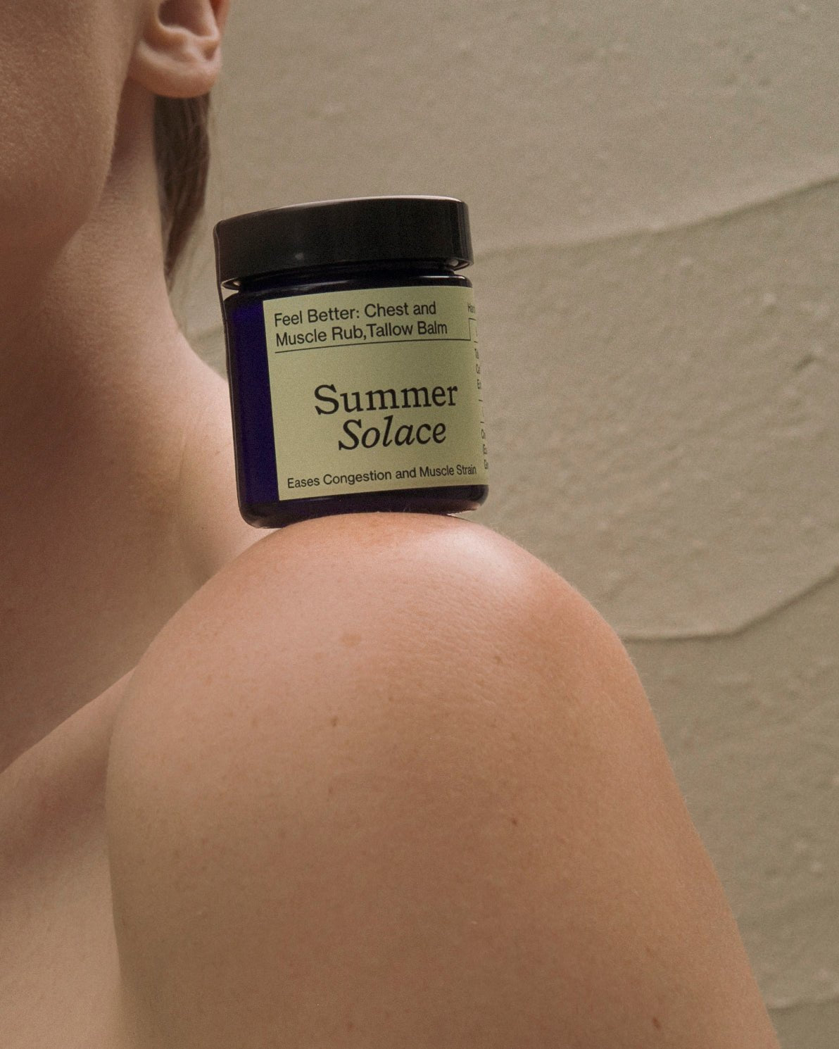 A small jar labeled "Summer Solace Tallow" balancing on a person's bare shoulder contains the product "Feel Better: Chest & Muscle Rub Balm - Regenerative Tallow™," designed to ease congestion and muscle strain. The person and background appear softly lit and muted.