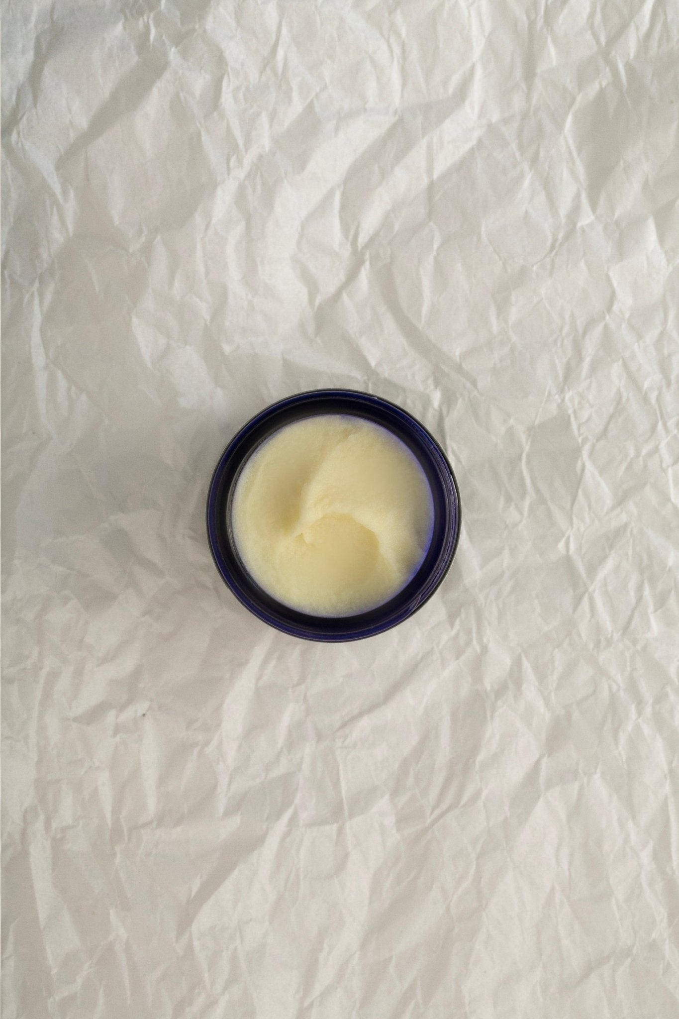 A small, open container with a blue lid sits on crumpled white paper. The container is filled with the Summer Solace Tallow Feel Better: Chest & Muscle Rub Balm - Regenerative Tallow™, showcasing a smooth, slightly swirled surface.