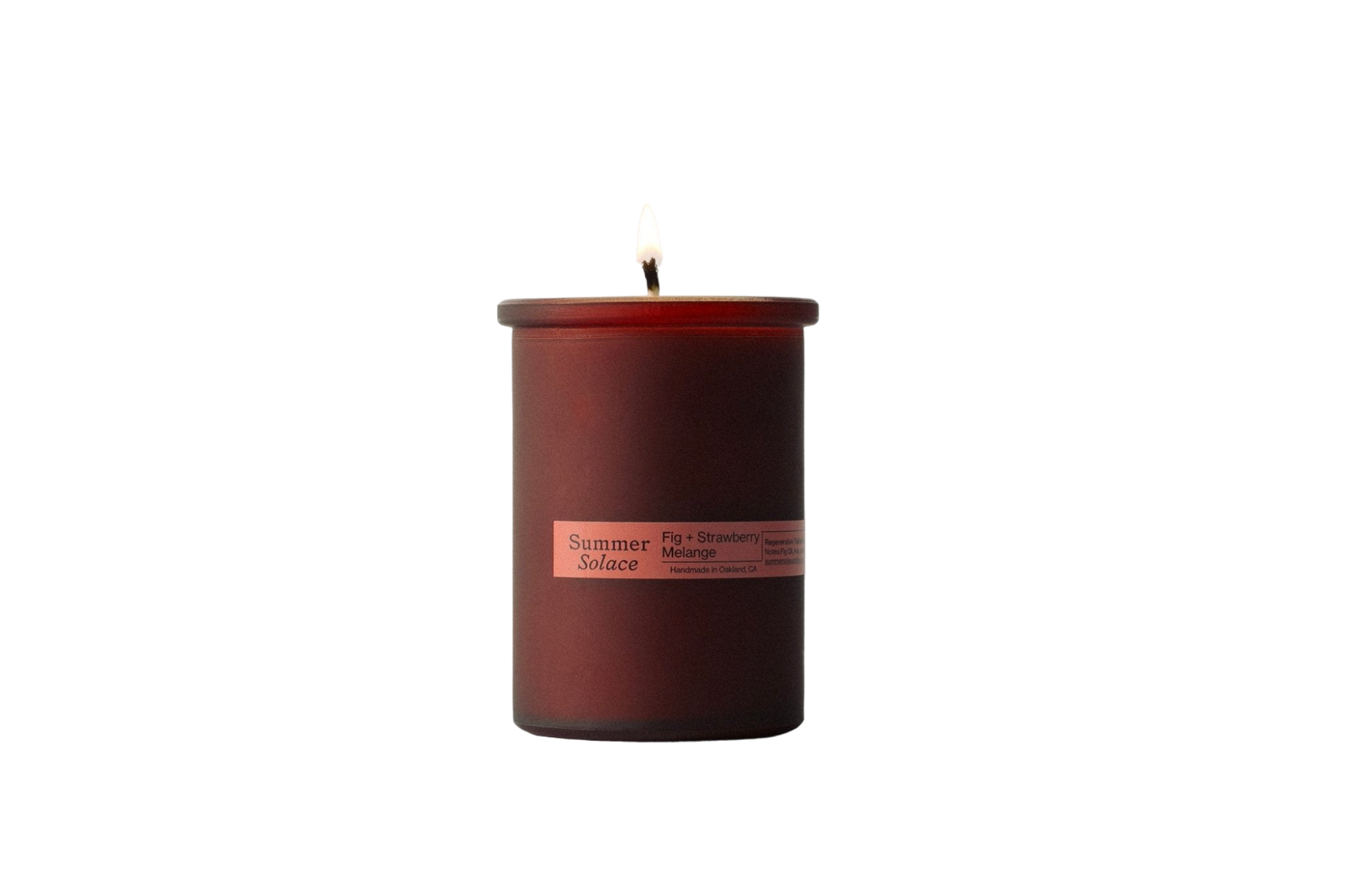 A single lit candle in a dark red cylindrical glass container is centered against a solid black background. The label on the Summer Solace Tallow candle reads "Fig and Strawberry Melange Candle - Regenerative Tallow™" and lists the scents "Fig, Strawberry, Melograno," highlighting its use of California fig leaf oil.