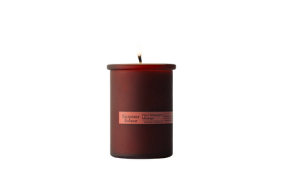 A single lit candle in a dark red cylindrical glass container is centered against a solid black background. The label on the Summer Solace Tallow candle reads "Fig and Strawberry Melange Candle - Regenerative Tallow™" and lists the scents "Fig, Strawberry, Melograno," highlighting its use of California fig leaf oil.