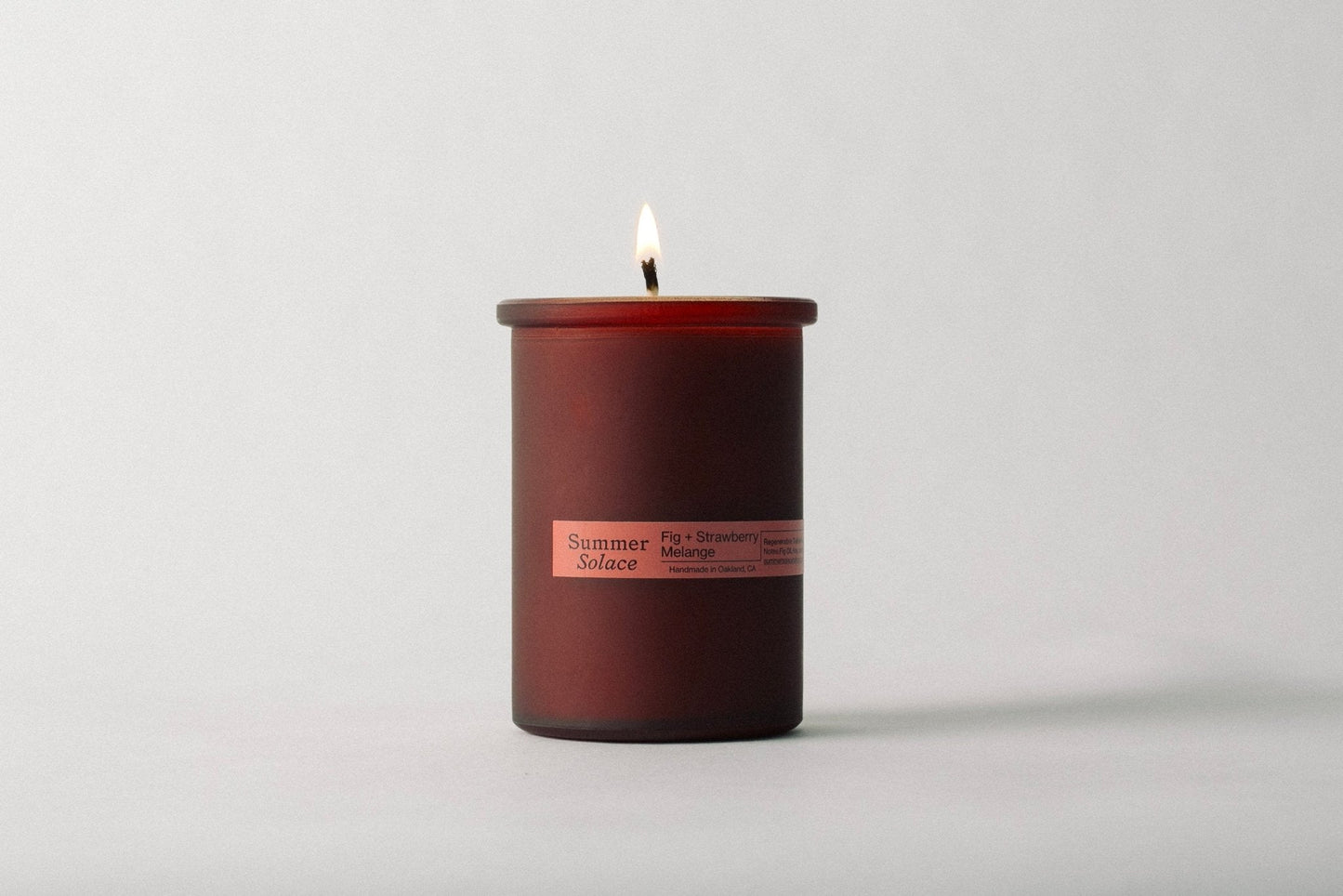 A Fig and Strawberry Melange Candle - Regenerative Tallow™ from Summer Solace Tallow is lit inside a cylindrical glass jar. The flame is visible against a plain, light-colored background, and the candle emits scents of fig, strawberry, and melon.