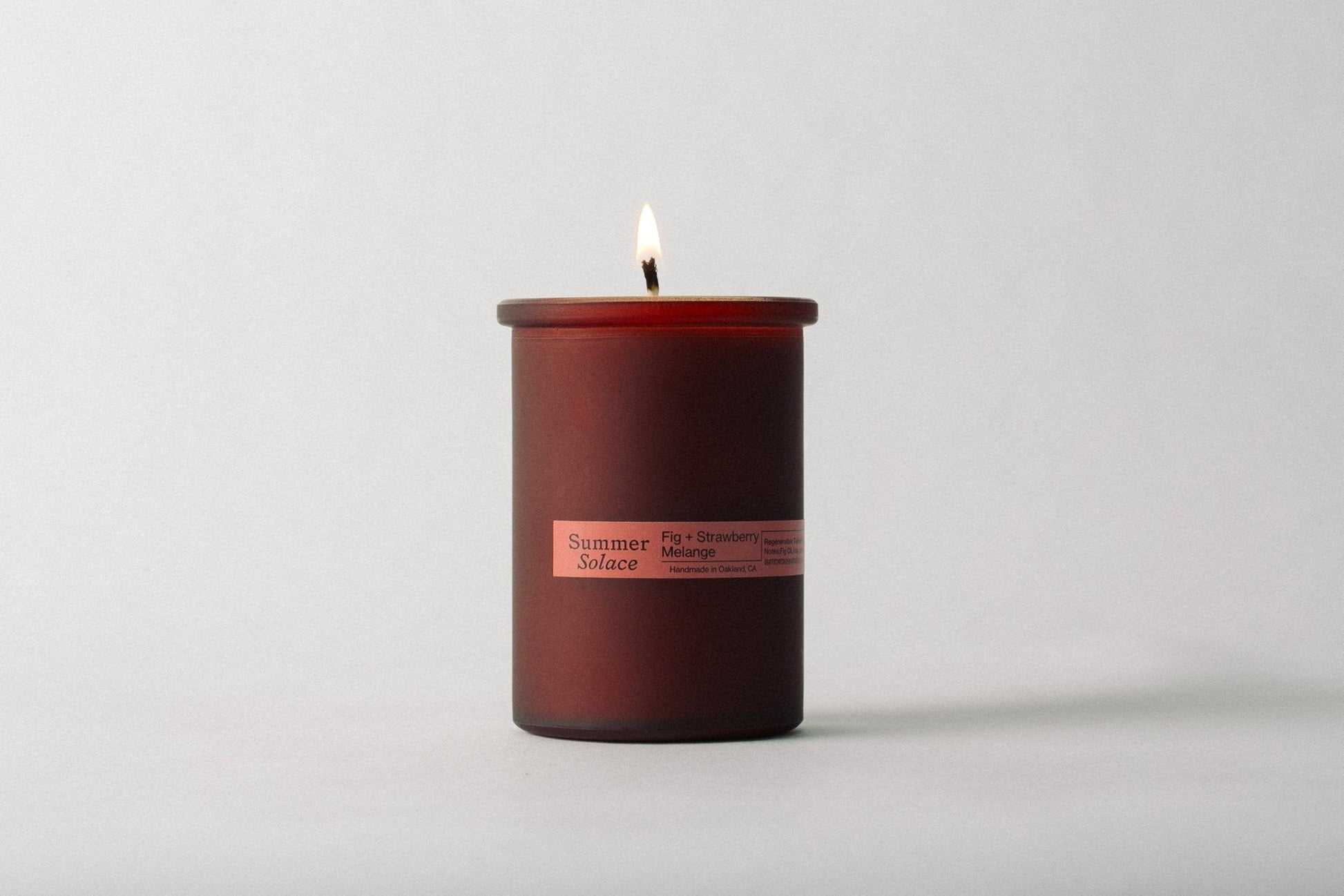 A Fig and Strawberry Melange Candle - Regenerative Tallow™ from Summer Solace Tallow is lit inside a cylindrical glass jar. The flame is visible against a plain, light-colored background, and the candle emits scents of fig, strawberry, and melon.