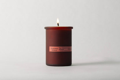 A Fig and Strawberry Melange Candle - Regenerative Tallow™ from Summer Solace Tallow is lit inside a cylindrical glass jar. The flame is visible against a plain, light-colored background, and the candle emits scents of fig, strawberry, and melon.