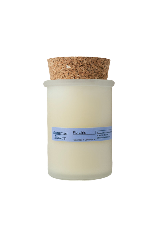 A frosted glass jar candle with a cork lid, featuring a light blue label that reads "Summer Solace Tallow" and "Flora Iris Candle - Regenerative Tallow™." It is handmade in Oakland, CA. The cream-colored wax ensures the sustainable tallow candles feel luxurious.