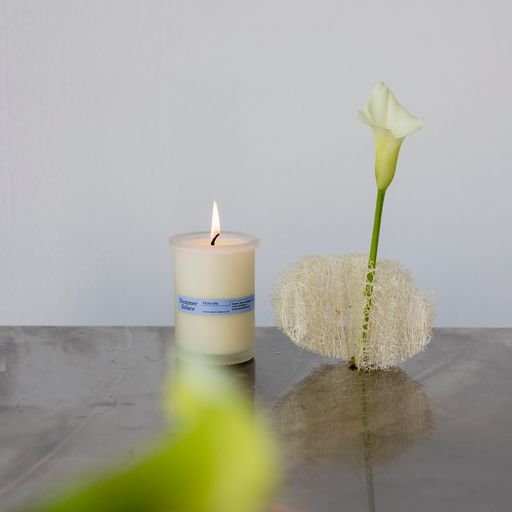 A lit Flora Iris Candle - Regenerative Tallow™ by Summer Solace Tallow in a minimalist container sits on a reflective gray surface next to a single green-stemmed white flower in a textured vase. The background is a plain, light-colored wall, creating an elegant yet simple ambiance.