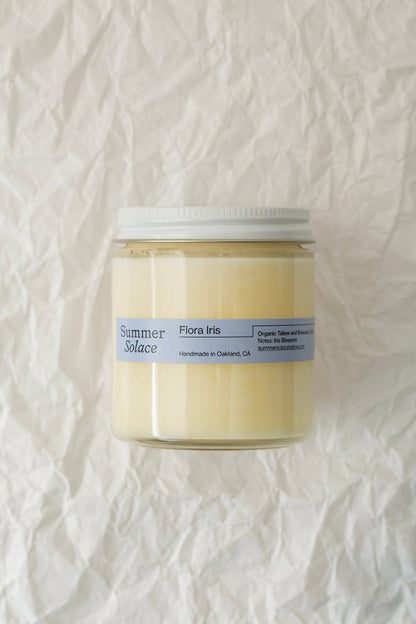 A glass jar tallow candle with a white lid set against a crumpled white background. The label reads "Summer Solace Tallow" and "Flora Iris Candle - Regenerative Tallow™," indicating it's handmade in Oakland, CA, and organic. With a pale yellow hue, this Flora Iris tallow candle includes the soothing scent of orris butter essential oil.