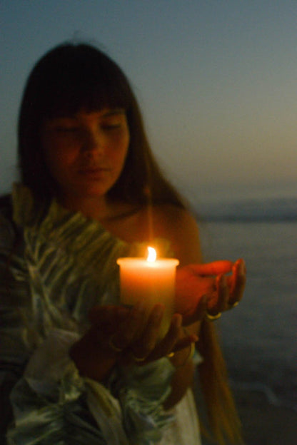 At dusk or dawn, a person with long hair stands near a body of water, holding a burning Flora Iris Candle - Regenerative Tallow™ from Summer Solace Tallow close to their face. They wear light-colored, textured clothing and appear contemplative as the sustainable candle illuminates their face in the low light.