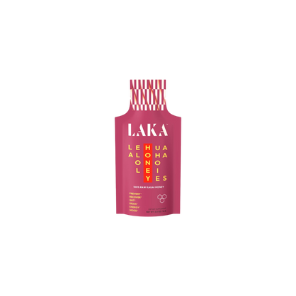 A pink packet labeled "LAKA" in white, featuring a yellow grid with the words "LEHUA HONEY ALOHA" displayed both horizontally and vertically. The text at the bottom states "100% RAW KAUAI HONEY," indicating it provides immune support with no added sugars or dairy, and promotes sweet energy. The cap is red and white striped. This product is known as LAKA Honey Sticks™ by the brand LAKA.
