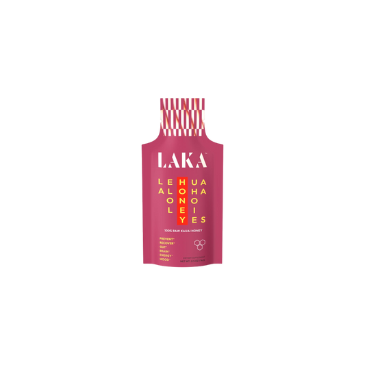 A pink packet labeled "LAKA" in white, featuring a yellow grid with the words "LEHUA HONEY ALOHA" displayed both horizontally and vertically. The text at the bottom states "100% RAW KAUAI HONEY," indicating it provides immune support with no added sugars or dairy, and promotes sweet energy. The cap is red and white striped. This product is known as LAKA Honey Sticks™ by the brand LAKA.
