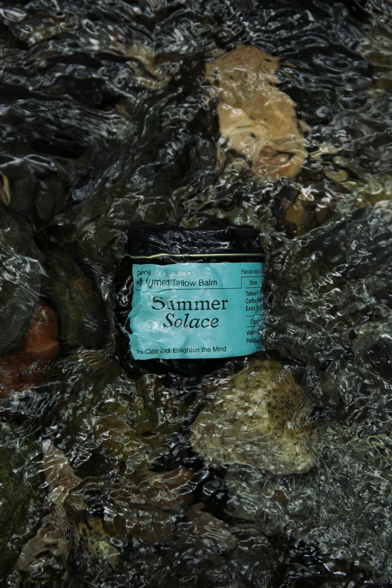 A jar of Summer Solace Tallow's Genji Perfumed Balm - Regenerative Tallow™ sits in a gently flowing river, surrounded by smooth stones and pebbles partially submerged in the clear water. The label on the jar is turquoise with black text, hinting at its woodsy scent and powerful moisturizing balm properties.