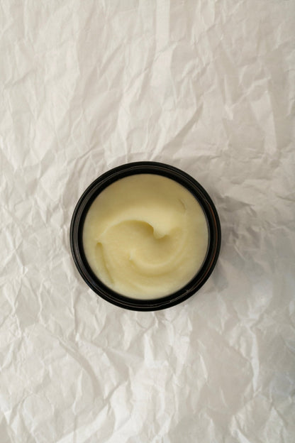 A dark container of Genji Perfumed Balm - Regenerative Tallow™ by Summer Solace Tallow is placed on a textured, crinkled white paper background. The creamy, light yellow balm within has a smooth surface with subtle swirls visible, hinting at its luxurious woodsy scent.