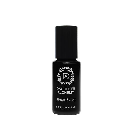 A small black bottle of Heart Salve® Botanical Nurturing Oil by Daughter Alchemy, crafted with organic ingredients, is displayed against a black background. The label features a white logo with a wreath design around the initials "DA" above the product name and indicates the bottle contains 0.3 fl oz (15 ml) of nurturing oil salve.